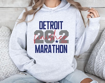 Op de afbeelding: Een lichtgrijze hoodie met de tekst "DETROIT MARATHON" in marineblauw, grijs en rood. De tekst bevat "26.2 Finisher 2025" in een gestileerd lettertype. De hoodie heeft een capuchon met trekkoord en lange mouwen.