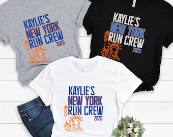 May include: Three t-shirts in gray, black, and white, each featuring the text "KAYLIE'S NEW YORK RUN CREW 2025" in blue and orange. The design includes an illustration of the Statue of Liberty. The shirts are short-sleeved.