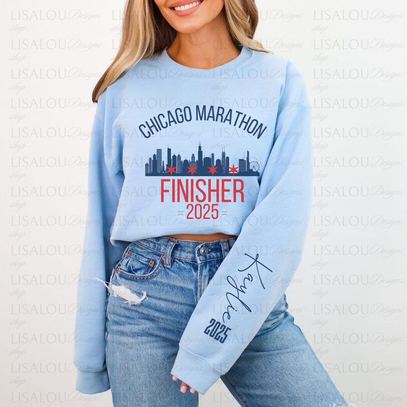 Op de afbeelding: Lichtblauw sweatshirt met de tekst "Chicago Marathon Finisher 2025" en een skyline-afbeelding. De mouw heeft de naam "Kaylie" en "2025" erop gedrukt. Een casual top voor marathonlopers.