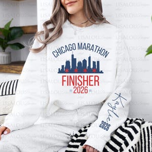 May include: A white crewneck sweatshirt featuring a navy blue Chicago Marathon skyline graphic with four red stars. The text reads Chicago Marathon, Finisher, and 2026 in red and blue. The left sleeve displays the name Kaylie and the year 2026 in navy blue.