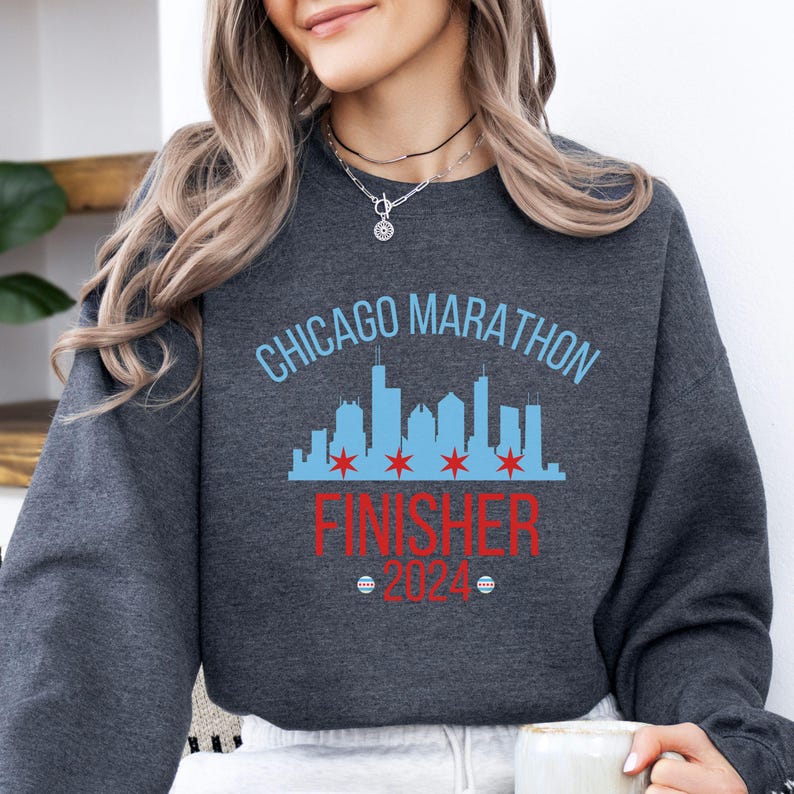 May include: A dark charcoal gray crewneck sweatshirt featuring a light blue Chicago skyline graphic with four red stars. The text reads CHICAGO MARATHON in light blue, FINISHER in red, and 2024 in red, flanked by two small circular Chicago flag icons.