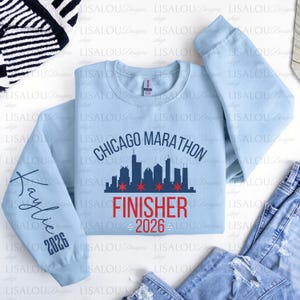 May include: A light blue long-sleeve crewneck sweatshirt featuring a dark blue Chicago Marathon skyline graphic with red stars. The text FINISHER 2026 is printed in red below the skyline. The left sleeve displays the name Kaylie and the year 2026.