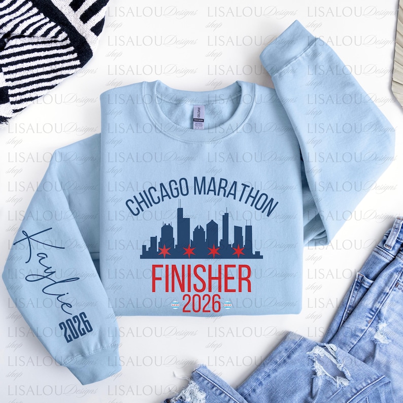 May include: A light blue long-sleeve crewneck sweatshirt featuring a dark blue Chicago Marathon skyline graphic with red stars. The text FINISHER 2026 is printed in red below the skyline. The left sleeve displays the name Kaylie and the year 2026.