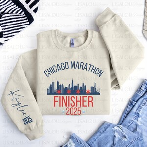 Op de afbeelding: Een beige Chicago Marathon Finisher sweatshirt met de tekst "Chicago Marathon" boven een skyline-afbeelding en "Finisher 2025" eronder. De mouw heeft de naam "Kaylie" en het jaar "2025".