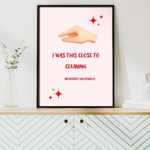 May include: Framed art print with a light pink background. The print features a hand emoji, the text "I WAS THIS CLOSE TO CLEANING" in red, and the quote "-DESPERATE HOUSEWIVES". Red starburst graphics are also included.
