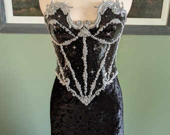 Pode incluir: Um vestido de veludo preto com um corpete preto e prateado. O corpete tem um design decorativo com detalhes prateados e estrelas.