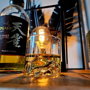 May include: A clear glass with a textured surface filled with amber liquid. The glass is sitting on a wooden surface next to a bottle of Tenjaku Pure Malt Whisky. The bottle is clear and has a black label with Japanese characters and the text "Tenjaku Whisky Pure Malt Product of Japan".