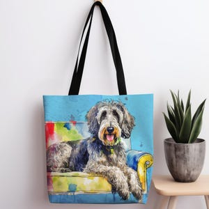 May include: A blue tote bag with a watercolor illustration of a dog sitting in a colorful armchair. The dog is gray and white with black fur and has a happy expression.