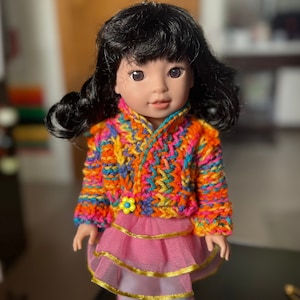 May include: A doll with black hair wearing a colorful crocheted sweater and a pink tutu.