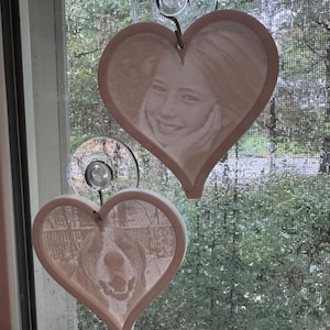 May include: Two white ceramic heart-shaped ornaments with suction cups. One ornament features a photo of a person smiling, and the other ornament features a photo of a dog.