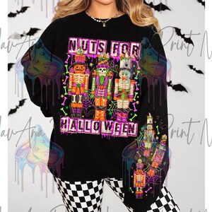 May include: Black long-sleeve shirt with the text "NUTS FOR HALLOWEEN" in a pink and purple font. The shirt features a graphic of three Halloween-themed nutcrackers in orange, purple, and green. The nutcrackers are decorated with spiderwebs and bones.