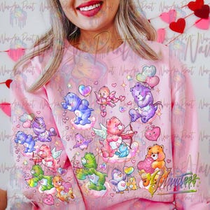 May include: Pink sweatshirt with a colorful print of cartoon bears with hearts and clouds. The bears are wearing wings and holding hearts and arrows.