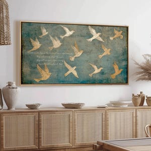 May include: A framed wall art piece featuring a teal background with gold-colored flying birds. The artwork has a gold frame and includes text. The piece is displayed above a wooden cabinet in a room setting.