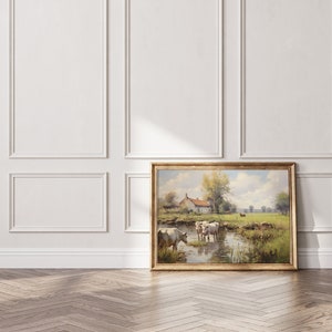 May include: A framed painting of a rural landscape with a farmhouse, trees, and cows wading in a stream. The painting is in a gold frame and is hanging on a white wall.
