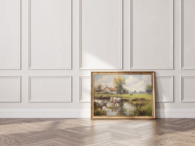 May include: A framed painting of a rural landscape with a farmhouse, trees, and cows wading in a stream. The painting is in a gold frame and is hanging on a white wall.