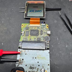 May include: Disassembled Game Boy console, showing internal components. The screen displays "GAME BOY" in blue. Circuit boards, wires, and a speaker are visible. A red probe is touching a circuit board. The console is being repaired.