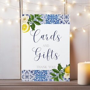 May include: A white sign with blue and white tile border and lemon and flower accents. The sign says "Cards and Gifts" with the text "Thank You!" below.