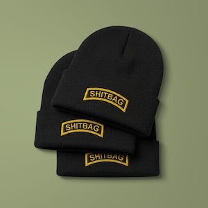 May include: A stack of three black knit beanies, each featuring a rectangular patch with a yellow border and the text SHITBAG embroidered in yellow capital letters. The beanies are made of a ribbed fabric and have a folded cuff design.