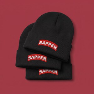 May include: Three black knit beanies stacked on top of each other. Each beanie features a rectangular red patch on the front cuff with the word SAPPER embroidered in white capital letters. The beanies have a ribbed texture and a folded brim.