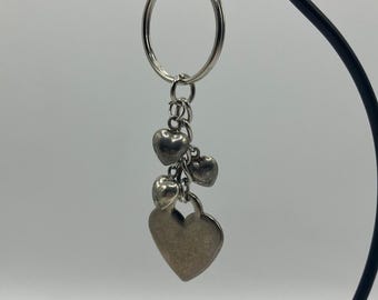 May include: Silver-toned keychain featuring a cluster of heart charms. The keychain has a large ring at the top, with a series of smaller hearts dangling below, culminating in a larger heart-shaped charm at the bottom. The hearts have a slightly textured surface.
