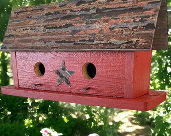 Könnte beinhalten: Ein rechteckiges Vogelhaus aus Holz mit leuchtend roter Lackierung und einem strukturierten, verwitterten Metalldach. Die Vorderseite hat zwei runde Einfluglöcher und einen mittigen Sternausschnitt aus Metall. Oben befindet sich eine Metallöse.