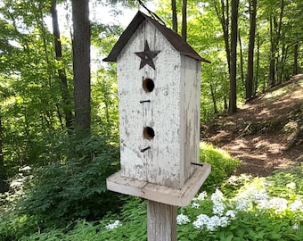 Könnte beinhalten: Verwittertes weißes Vogelhaus mit rustikaler Ästhetik. Es hat ein braunes Metalldach, einen Sternakzent und zwei kreisförmige Eingangsöffnungen. Das Vogelhaus ist auf einem Holzpfosten montiert, umgeben von üppigem Grün und Bäumen.