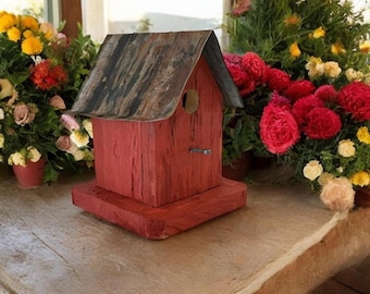 May include: A red wooden birdhouse featuring a weathered, dark gray metal roof with a peaked design. The structure has a single circular entrance hole on the front facade and a small horizontal perch extending from the center of the red wood wall.