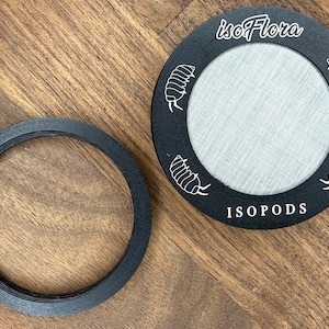 Puede incluir: Dos objetos circulares negros sobre una superficie de madera. Uno es un anillo sólido, el otro es un marco circular con un centro de malla y el texto "isoFlora" e "ISOPODS". El marco tiene ilustraciones de insectos blancos.