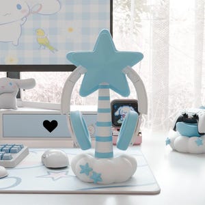 Puede incluir: Un soporte para auriculares en forma de estrella azul y blanco con una base en forma de nube. El soporte está diseñado para sostener los auriculares y tiene un poste a rayas azul y blanco. El soporte está sobre un escritorio blanco con un teclado y un ratón azul y blanco.