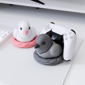 Puede incluir: Una figura de pájaro blanco con pico rosa en un nido rosa, y una figura de pájaro gris con pico negro en un nido gris, que sirve de soporte para un mando de juego inalámbrico blanco y negro con el logotipo de PlayStation.