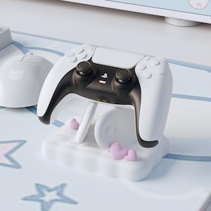 May include: A white and black PlayStation 5 controller on a white cloud-shaped charging stand. The stand features three small pink heart accents. A white computer mouse is visible in the background.