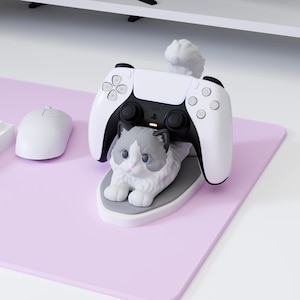 May include: A white and gray cat-shaped controller stand holding a white and black game controller. The cat figure has a fluffy tail, blue eyes, and a pink nose, resting on a gray oval base. A white computer mouse is positioned to the left.