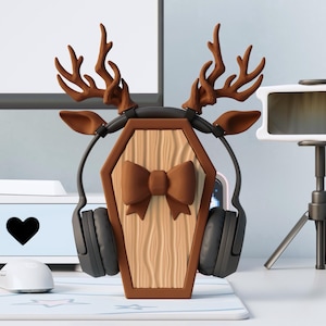 Può includere: Un supporto per cuffie marrone e beige a forma di bara con un papillon, corna e orecchie di cervo. Il supporto contiene un paio di cuffie nere. Il supporto è su una scrivania con un monitor del computer e altri accessori sullo sfondo.