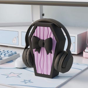 Könnte beinhalten: Schwarzer Kopfhörer auf einem schwarzen Sarg-Ständer mit rosa Holzmaserung und schwarzer Schleife. Der Ständer steht auf einem Schreibtisch mit Tastatur und Maus.