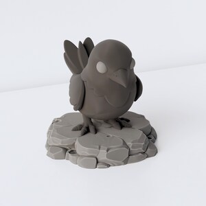 May include: A small, matte dark grey figurine of a bird with large, rounded eyes and a pointed beak. The bird stands on a textured, circular base that resembles a cluster of uneven, flat stones, all rendered in a uniform dark grey colour.