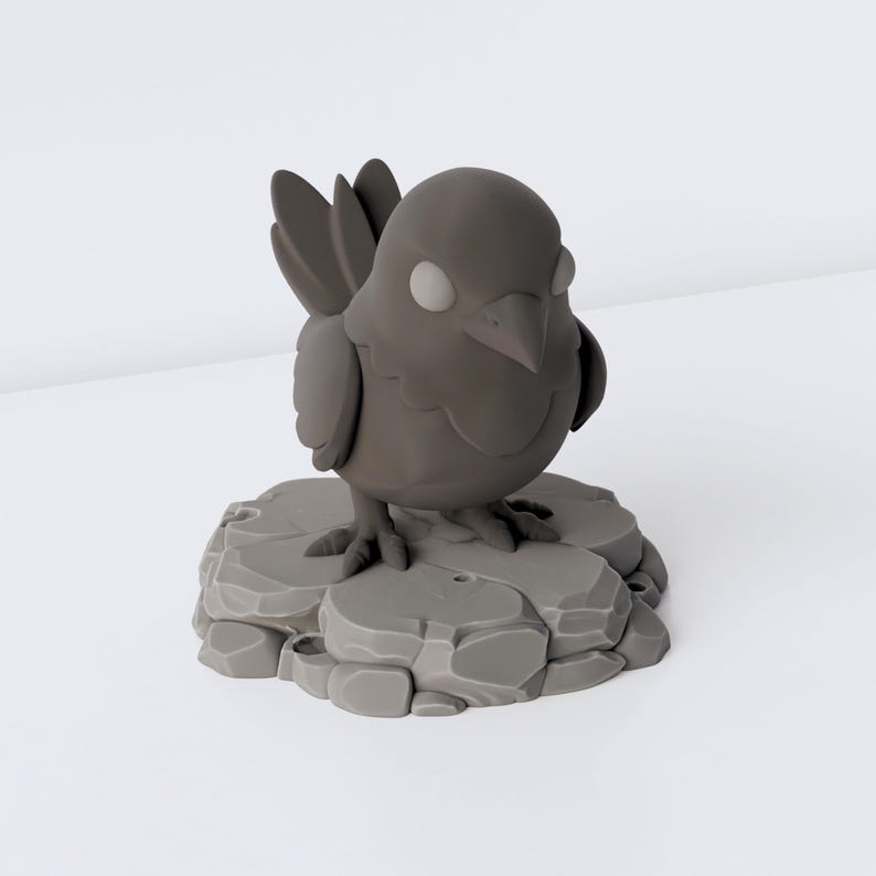 May include: A small, matte dark grey figurine of a bird with large, rounded eyes and a pointed beak. The bird stands on a textured, circular base that resembles a cluster of uneven, flat stones, all rendered in a uniform dark grey colour.