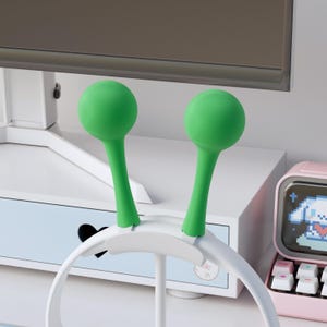 Può includere: Un supporto per cuffie bianco con due elementi sferici verdi. Il supporto è su una superficie bianca, con un cassetto azzurro e un monitor per computer rosa in stile retrò sullo sfondo.