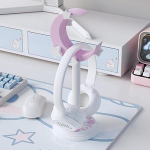 Peut inclure: Un support de casque blanc et violet pastel avec un motif de croissant de lune et d'étoiles au sommet, soutenu par un pilier vertical cannelé sur une base circulaire avec des accents ronds, portant un casque audio blanc supra-auriculaire.