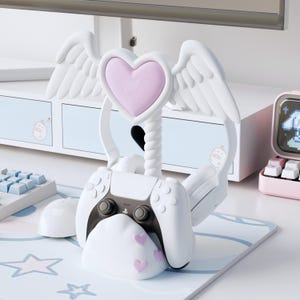 May include: White gaming controller stand with a heart and wing design. The stand holds a white and black controller, and features a pink heart and wings. The base has pink hearts. A gaming mouse and keyboard are in the background.