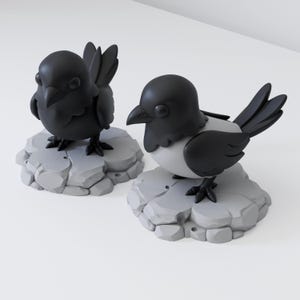 May include: Two stylised bird figurines featuring matte black heads, wings, and tails with light grey chests. Each bird stands on a circular, light grey base textured to resemble a cluster of smooth, rounded stones or rocks on a plain surface.