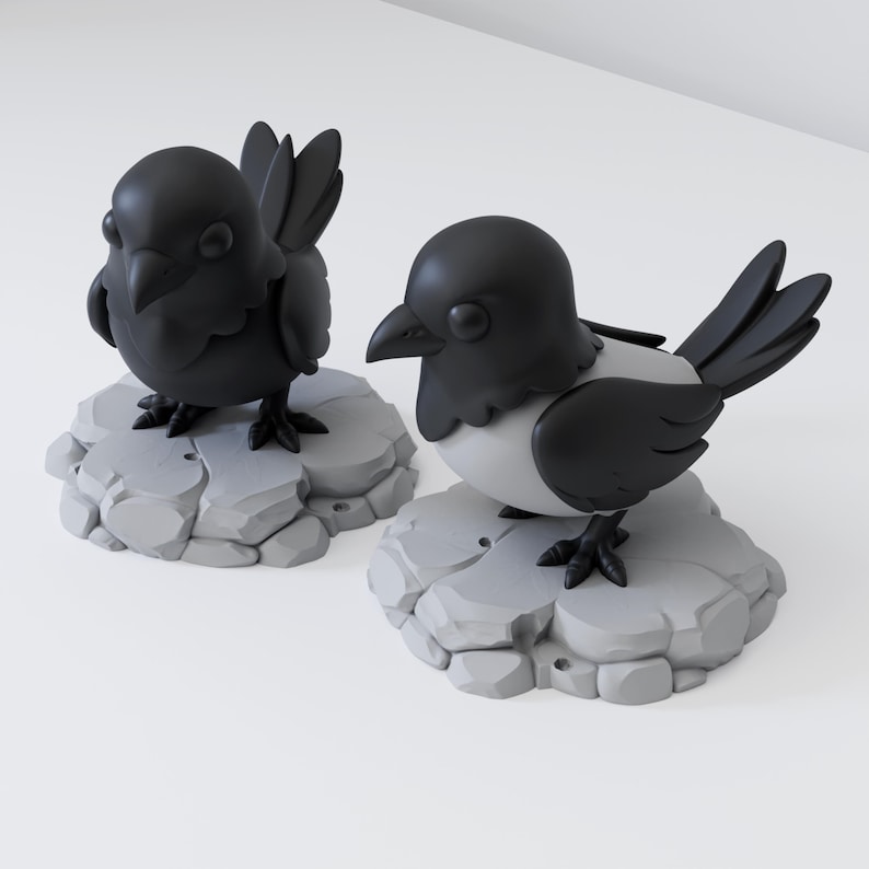 May include: Two stylised bird figurines featuring matte black heads, wings, and tails with light grey chests. Each bird stands on a circular, light grey base textured to resemble a cluster of smooth, rounded stones or rocks on a plain surface.