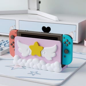 Può includere: Un supporto di ricarica rosa per Nintendo Switch con una stella gialla e ali bianche. Il supporto ha una base a forma di nuvola e tiene la console Switch con Joy-Con corallo e turchese. Lo sfondo include una scrivania e un cassetto con maniglia a forma di cuore.