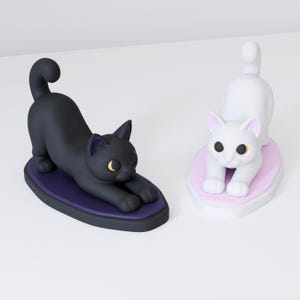 May include: Two cat figurines in a stretching pose. One is a solid black cat on a dark purple oval base with yellow eyes. The other is a solid white cat on a light pink oval base with yellow eyes. Both cats have arched backs and tails raised.