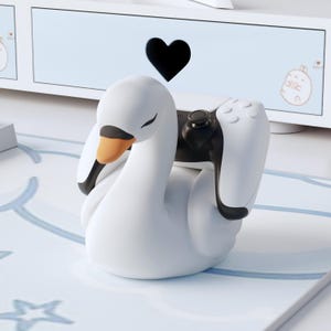 May include: A white swan-shaped controller holder with a black and orange beak, holding a black and white game controller. The holder is on a light blue surface with a heart-shaped cutout in the background.