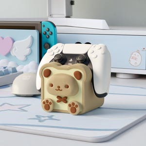 Può includere: Un supporto per controller beige e marrone a forma di fetta di pane con la faccia di un orso. Un controller PlayStation bianco è inserito nel supporto. Il supporto è su un tappetino da scrivania blu e bianco.