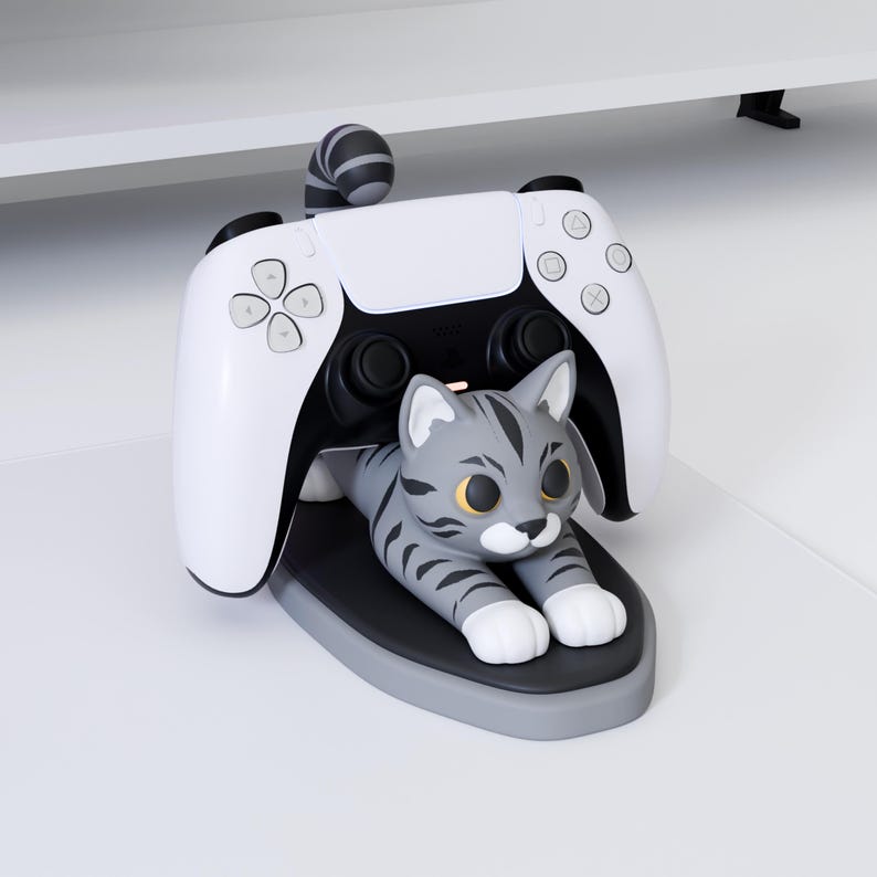 May include: A white and black video game controller resting on a gray oval stand featuring a sculpted gray tabby cat with dark stripes, large yellow eyes, and white paws. The cat's tail is curled upward behind the controller's central section.