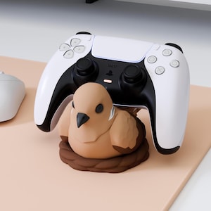 Könnte beinhalten: Ein weiß-schwarzer kabelloser Videospiel-Controller auf einem beigen, vogelgeformten Ständer. Die Vogelfigur ist hellbraun mit einem dunkelbraunen Schnabel und schwarzen Augen und sitzt auf einer dunkelbraunen, kreisförmigen Nestbasis.