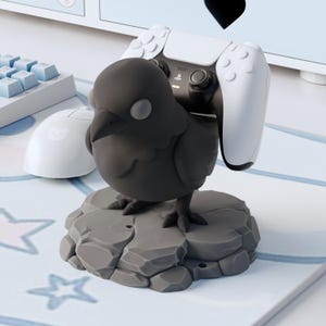 May include: A matte black bird figurine controller stand perched on a gray rock-textured base. The bird is designed to hold a white and black gaming controller on its back. The figure features a smooth, rounded body with a pointed beak and eyes.
