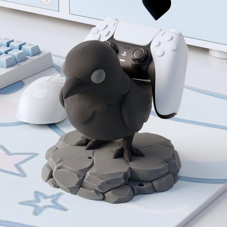 May include: A matte black bird figurine controller stand perched on a grey rock-textured base. The bird is designed to hold a white and black gaming controller on its back. The figure features a smooth, rounded body with a pointed beak and eyes.