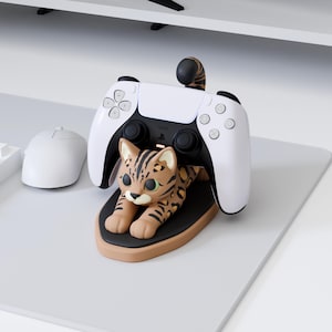 Op de afbeelding: Een gaming controller standaard met een bruin en zwart gevlekte luipaardfiguur in liggende positie. De standaard is ontworpen om een witte en zwarte draadloze gaming controller vast te houden, waarbij de staart van de luipaard achter de controller omhoog steekt.